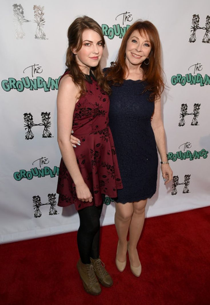 two women, one younger and one older, stand on the red carpet at a premiere event. the younger woman is wearing a dark maroon floral dress with black tights and black boots. the older woman is wearing a navy blue lace dress with nude heels. they are both smiling and the younger woman has her arm around the older woman's waist. they appear to be mother and daughter.  the words "the groundlings" is repeated on the backdrop.
