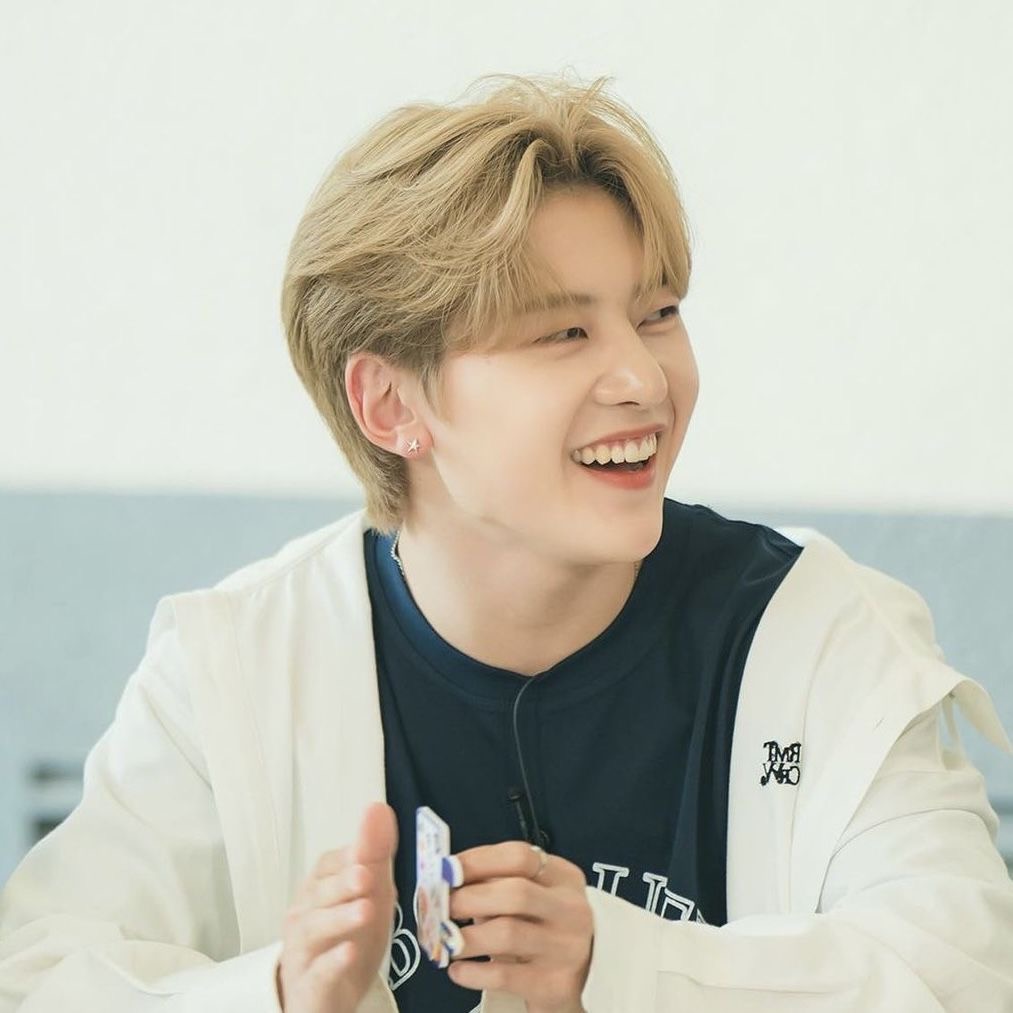 a portrait of a young asian man with blond hair smiling while holding a small object. he is wearing a white overshirt and a navy blue tshirt.  the background is a solid light gray.