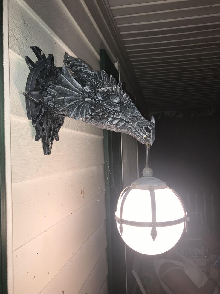 a gray dragon head sculpture is mounted on a white siding wall, with a round, glowing light fixture hanging from its mouth. the dragon is intricately detailed with scales, horns, and wings, giving it a fearsome appearance. the light fixture is a globe shape with metal supports, adding a decorative element to the scene. the overall impression is that of a fantasythemed outdoor decoration on a house.