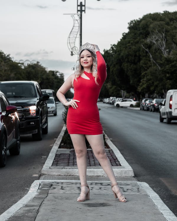 a fullbody shot of a young woman wearing a red oneshoulder dress and a crown, posing confidently on a city sidewalk. she is standing in front of a street with cars parked on both sides. the background shows lush greenery and a partly cloudy sky. the woman's expression is happy and she is looking directly at the camera.  the overall mood is celebratory and joyful, suggesting a beauty pageant or similar event.