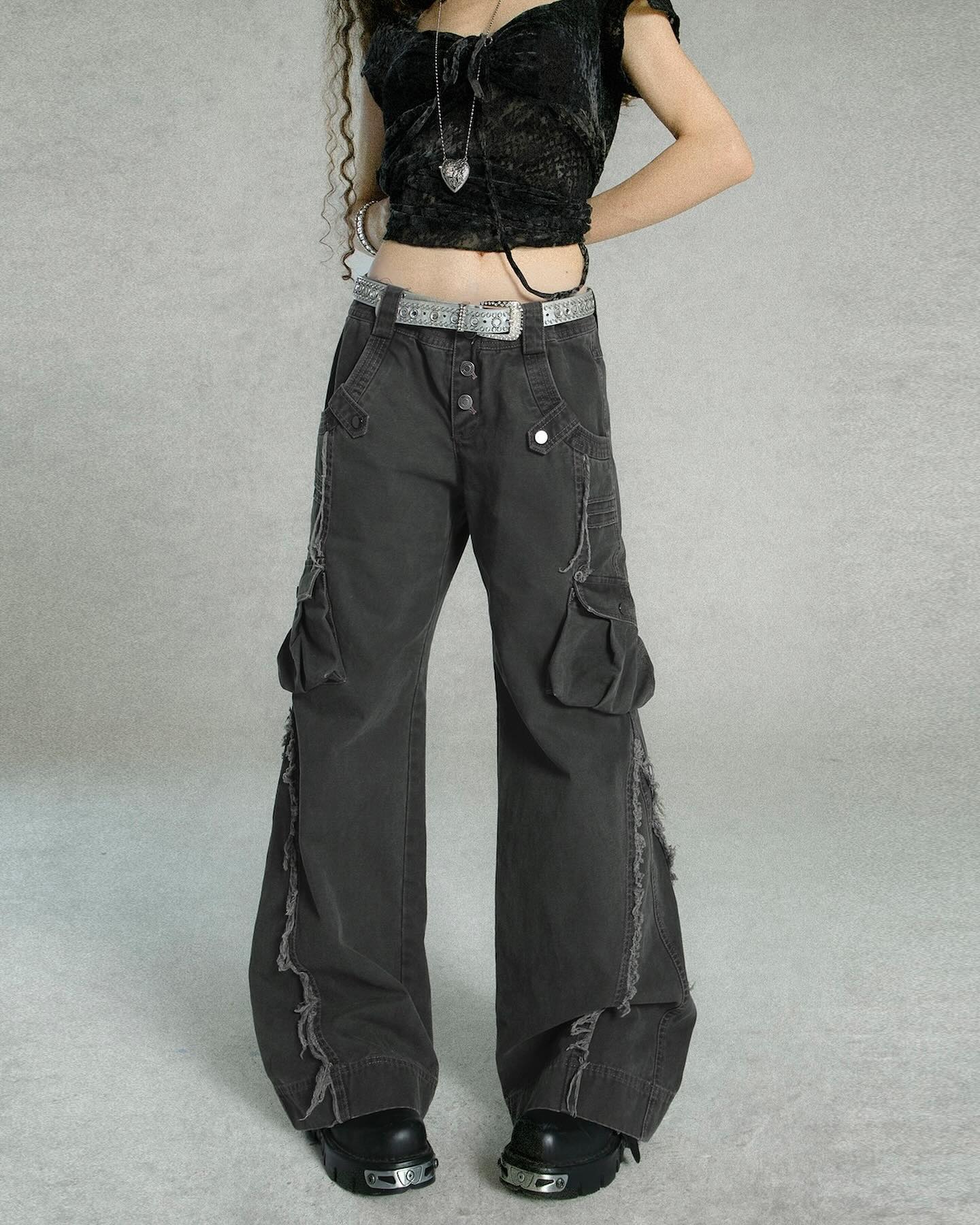 a full shot of a model wearing dark gray cargo pants with distressed details. the pants are widelegged, lowrise, and have visible fraying along the sides. the model is also wearing a black crop top, silver belt, and black boots. the overall style is grunge, alternative, and reminiscent of y2k fashion. the background is a plain gray wall. the image is welllit and showcases the details of the clothing.