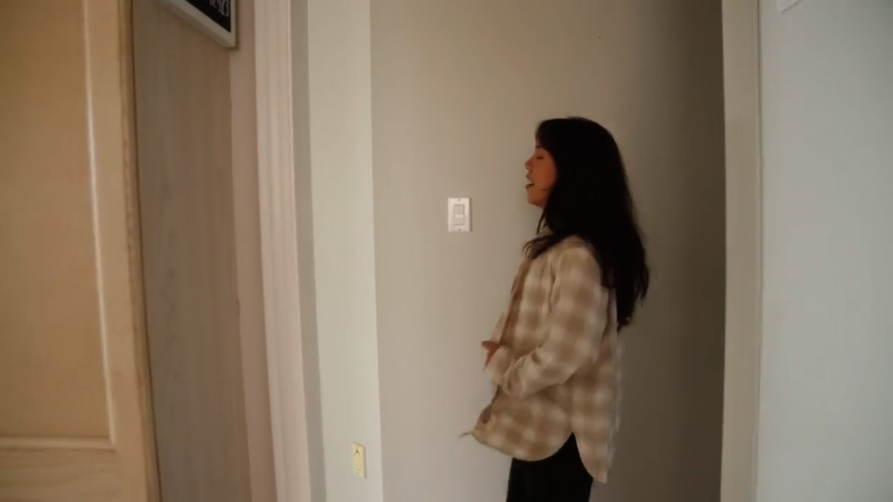 woman with long dark hair, wearing a beige and white plaid shirt and black pants, is standing in a hallway. the hallway is painted a light gray, and the trim is white. there is a door on the left side of the image, and a doorway on the right side. the woman is facing the left side of the image, and her mouth is slightly open as if she is talking. the lighting in the image is bright and natural, coming from the doorway on the left side of the image.