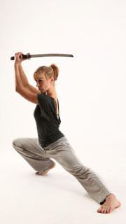 a woman with her hair pulled back in a ponytail is holding a katana over her head. she is wearing grey yoga pants and a charcoal grey shirt. she is barefoot and appears to be in an attack position. the background is a plain white.  the lighting is soft.