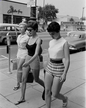 a vintage black and white photograph shows three stylish women walking down a sidewalk on a sunny day in the 1960s. the women are dressed in fashionable shorts and tops. the woman on the left wears a collared shirt, belted shorts, and carries a woven bag. the woman in the middle wears a black top, checkered shorts, sunglasses, and carries a purse. the woman on the right wears a crop top and patterned shorts. all three wear sandals. a vintage car is parked along the curb, and a building with a sign is visible in the background. a parking meter stands on the sidewalk next to the women. the overall scene evokes a sense of midcentury style and leisure.