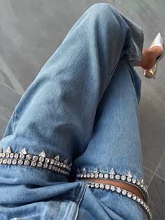 a closeup shot of a person wearing a pair of highwaisted, lightwash denim jeans with two rhinestone bands on the waistband. the jeans are fitted and the model's legs are slightly crossed, highlighting the sparkly embellishment. the model is wearing silver shoes with a pointed toe, adding to the stylish and contemporary look. the image is likely from a fashion shoot and the jeans are the main focus, showcasing their detail and texture. the background is blurred, drawing the viewer's attention to the jeans.