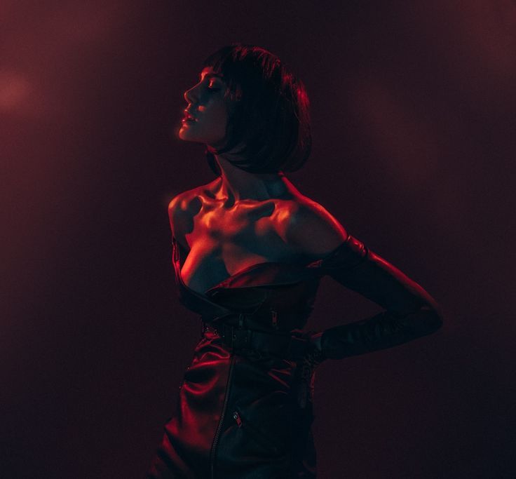 a woman in a black leather dress and gloves poses dramatically against a dark background lit with red light. her short black hair is styled in a sleek bob, and her expression is intense and suggestive. the image is moody and mysterious, with a strong emphasis on the woman's body and the luxurious textures of her outfit.
