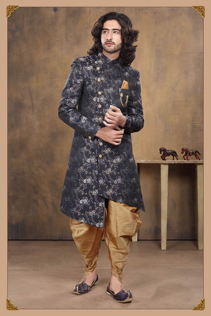 the image showcases a male model dressed in a stylish indowestern outfit. he is wearing a navy blue jacquard kurta with a floral print, designed as a short sherwani style with an asymmetrical hemline.  it is paired with gold dhotistyle pants. the kurta features button detailing and a small gold pocket square. he is also wearing dark blue patterned juttis (traditional indian footwear). the backdrop is a muted brown studio setting with small wooden horses visible on a shelf in the background. the overall look blends traditional indian elements with a modern, contemporary design.