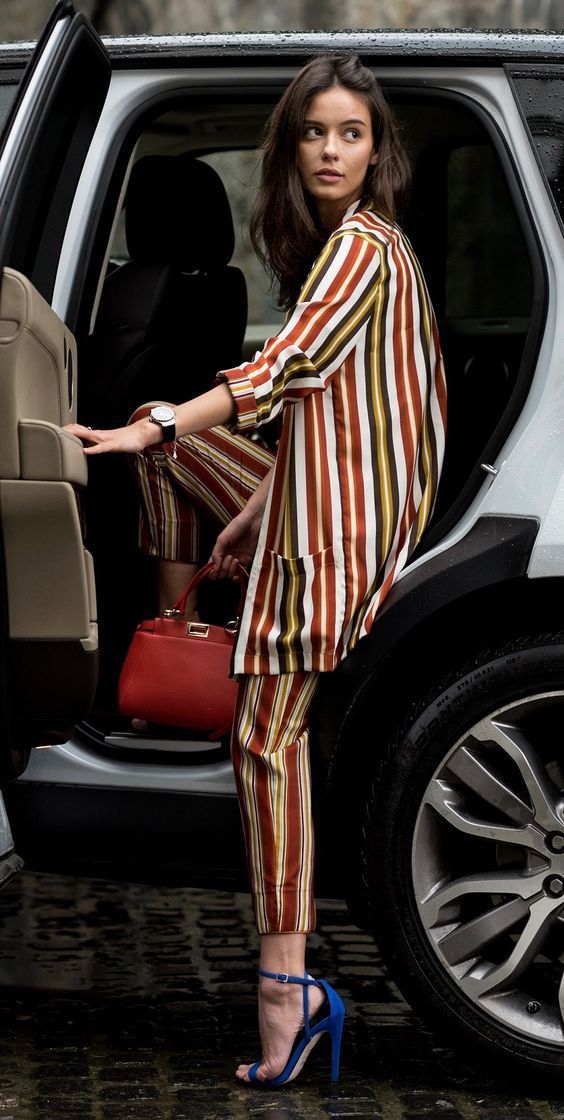 a stylish woman in a red and gold striped pantsuit and blue heels is getting out of a luxury range rover. she carries a red handbag. the scene is set in a city street. the woman looks elegant and confident.