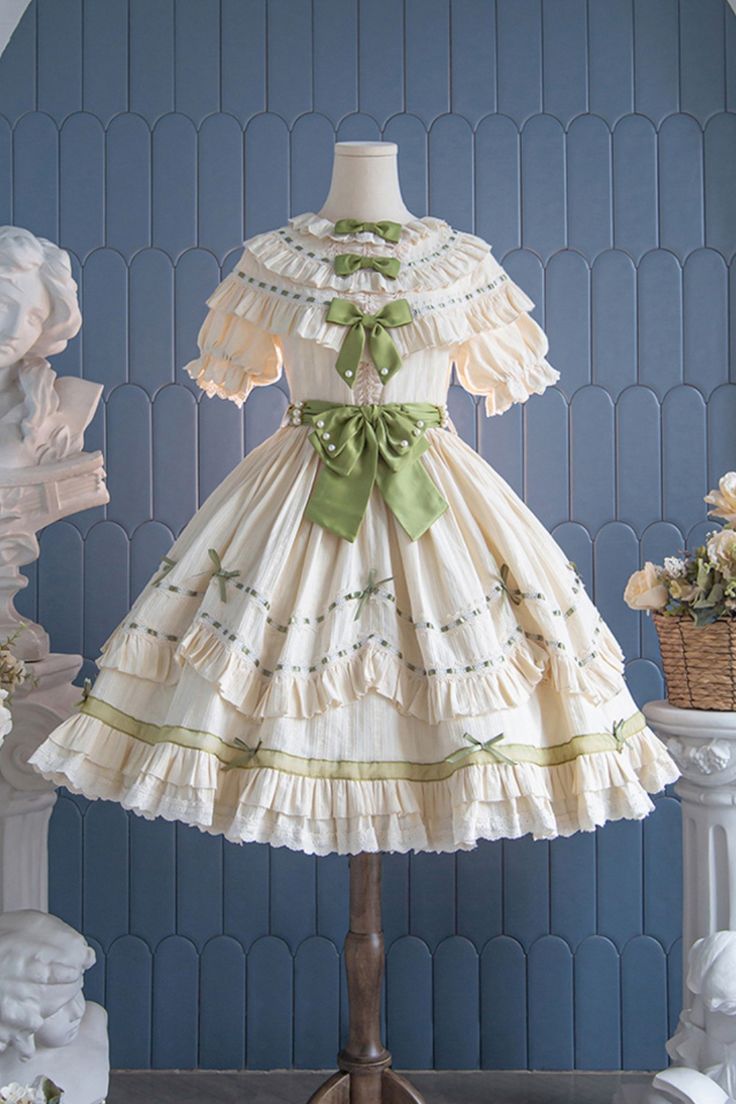 a creamy white lolita fashion dress is displayed on a mannequin in front of a bluetiled wall. the dress features intricate lace, ruffles, and is adorned with green ribbon accents. the soft lighting highlights the delicate details of the garment and creates a dreamy atmosphere.