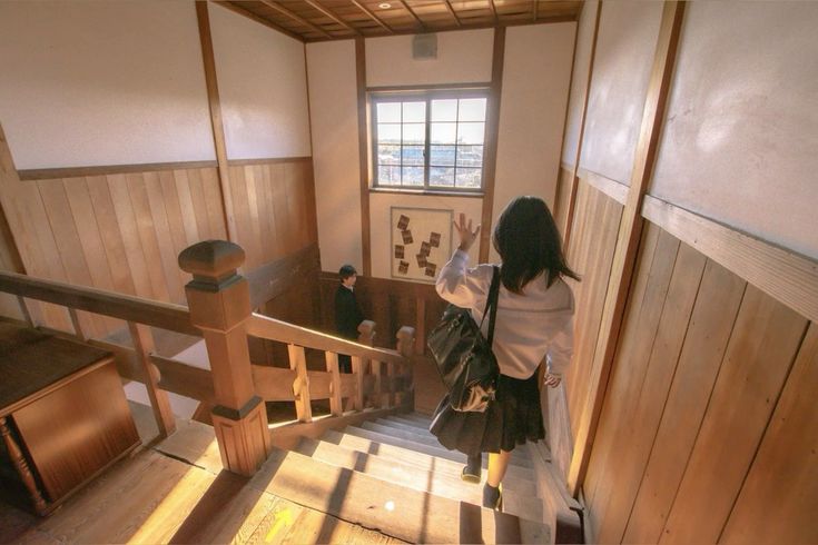 an image of a vintage school hallway, with wooden walls, floors, and stairs. there's a girl in a school uniform standing on the steps, waving her hand. a boy in a school uniform is standing at the top of the stairs and watching her.  the window is letting in a bright light from the sunshine.