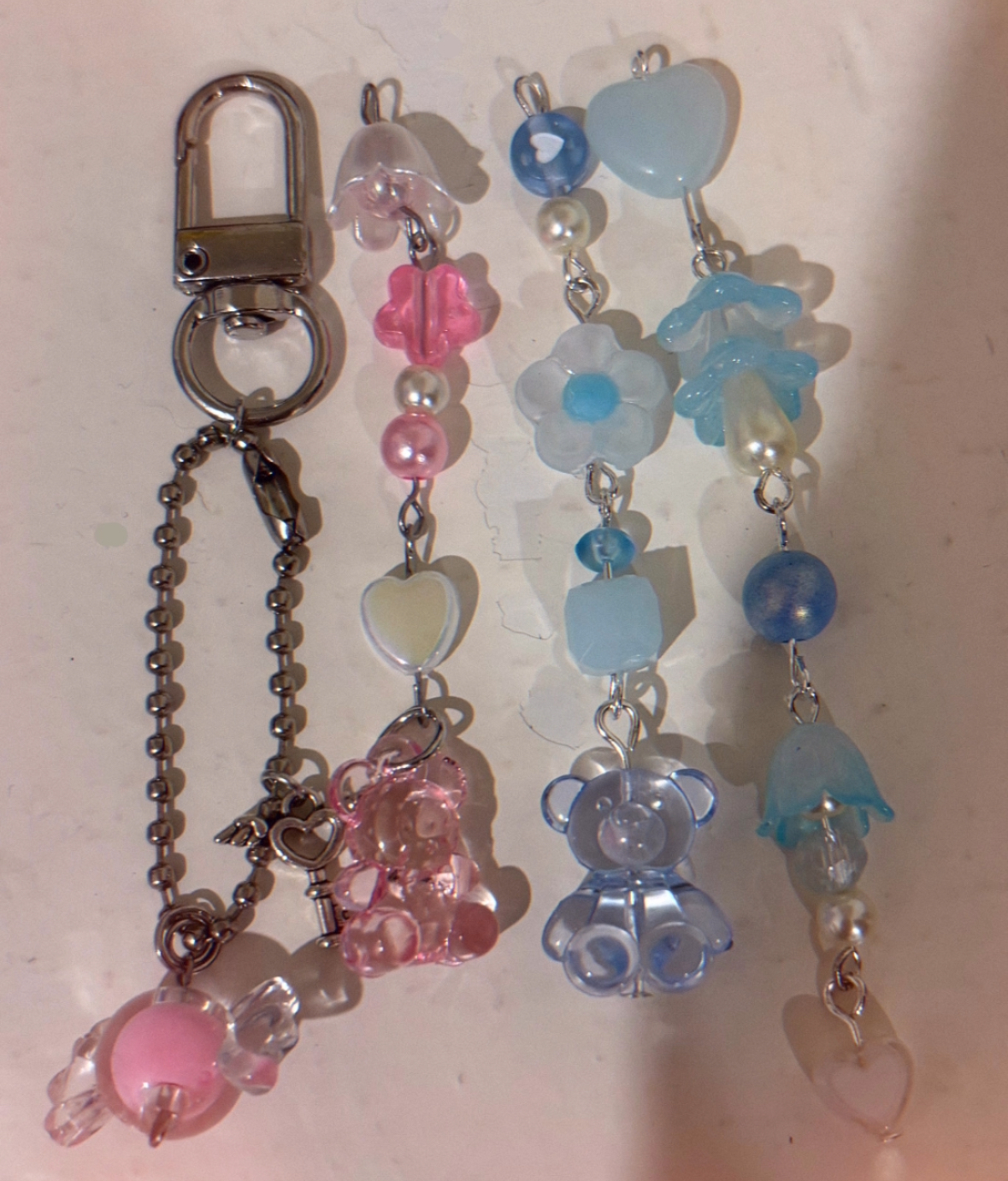 the image shows four handmade keychains featuring pastelcolored beads and charms. the leftmost keychain has a silver swivel clasp, a ball chain, a heart and winged key charm, and a pink candy charm. the next keychain has a pink flower, pearl beads, and a white heart, with a pink bear charm at the bottom. two other keychains are made of blue beads, flowers, and bears. these have silver connecting loops.