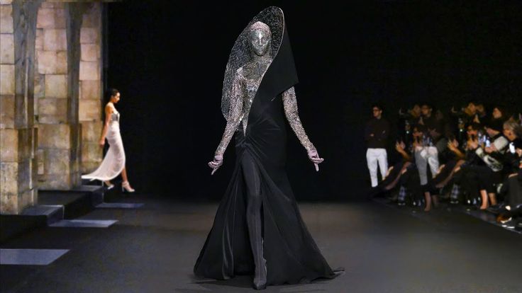 a fashion show scene featuring a model in a striking black and silver haute couture dress with a distinctive metallic veil. the model is standing on a runway, posing with her arms slightly extended. the dress has a dramatic silhouette, with a fitted bodice and a long, flowing skirt. the veil covers her head and face, creating a mysterious and futuristic look. in the background, an audience watches the show, some holding up phones to take pictures. to the left, another model walks down stairs next to a stone wall.