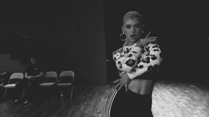 a black and white image of a woman with blonde hair wearing a leopard print sweater and black pants. she is wearing hoop earrings and appears to be in a dance studio. in the background, a man is sitting in a chair.