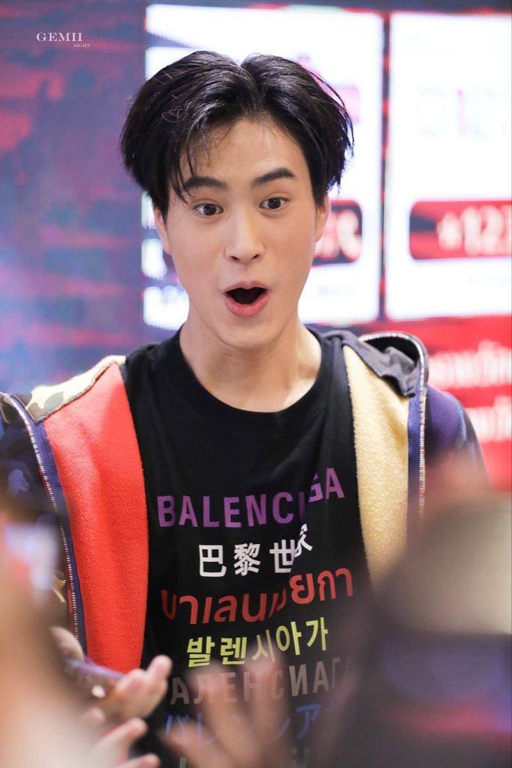 a man with short black hair and surprised expression is looking to his left. he's wearing a black balenciaga tshirt with a red and white design and a navy blue jacket with a camouflage design. the background is blurry and features a digital screen and some people.