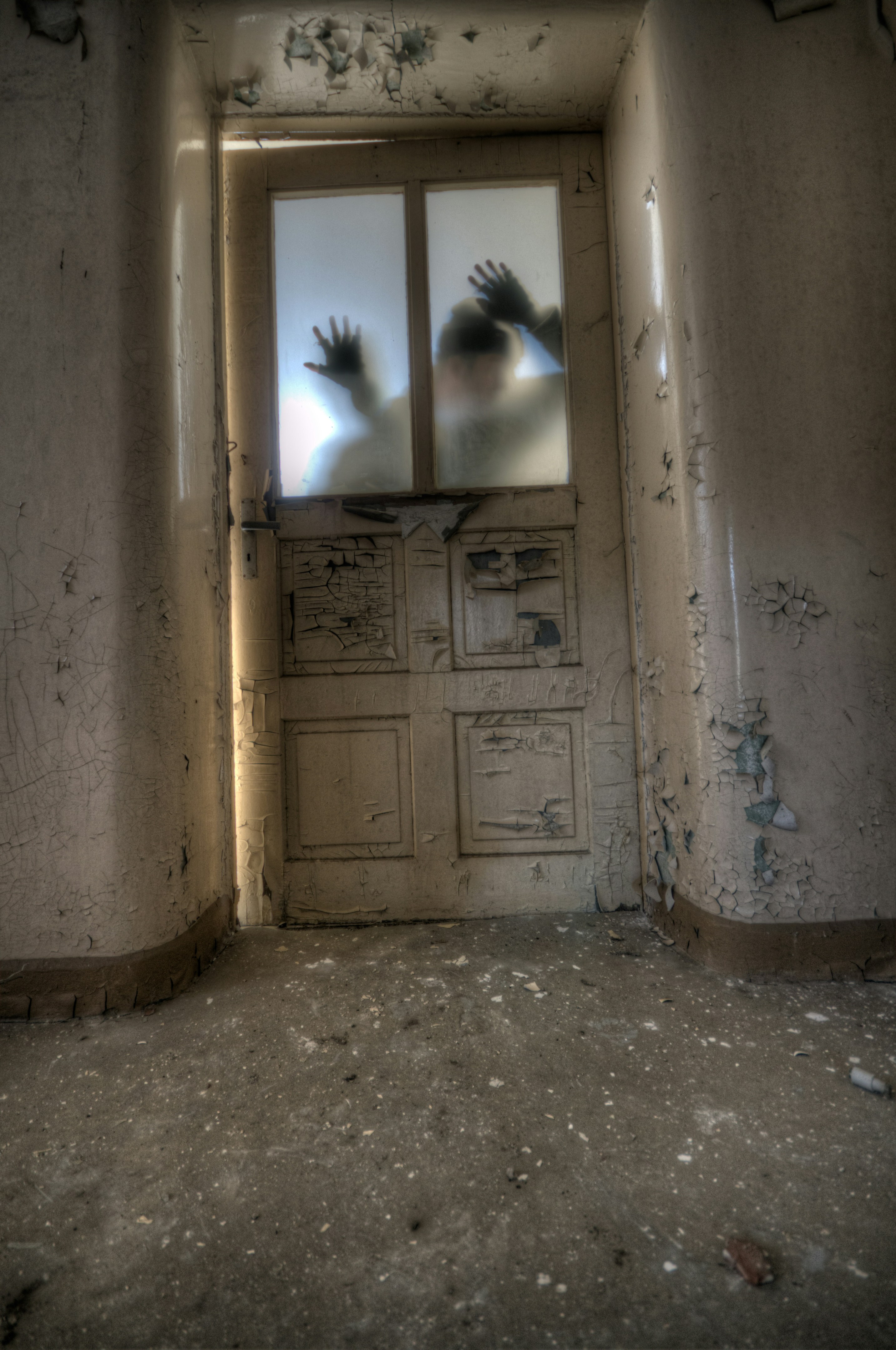 a worn and weathered door, its oncewhite paint now peeling and discolored, stands slightly ajar at the end of a narrow hallway.  the room beyond is dark, but a shadowy figure with raised hands is faintly visible through the frosted glass panes of the upper portion. the walls of the hallway are similarly dilapidated, hinting at years of neglect. the overall impression is one of unease and abandonment, evocative of haunted spaces.