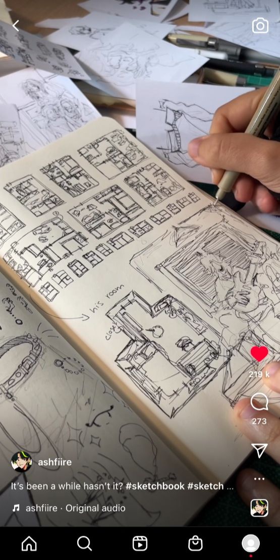 a close up image of a sketchbook with black ink drawings. the top of the sketchbook is partially out of the frame as a hand is holding a pen that is resting on the page. in the bottom right hand corner of the page a person is in the reflection of the sketchbook. there are many details on the page, the image is focused on the bottom half of the page, where a more detailed drawing is made.