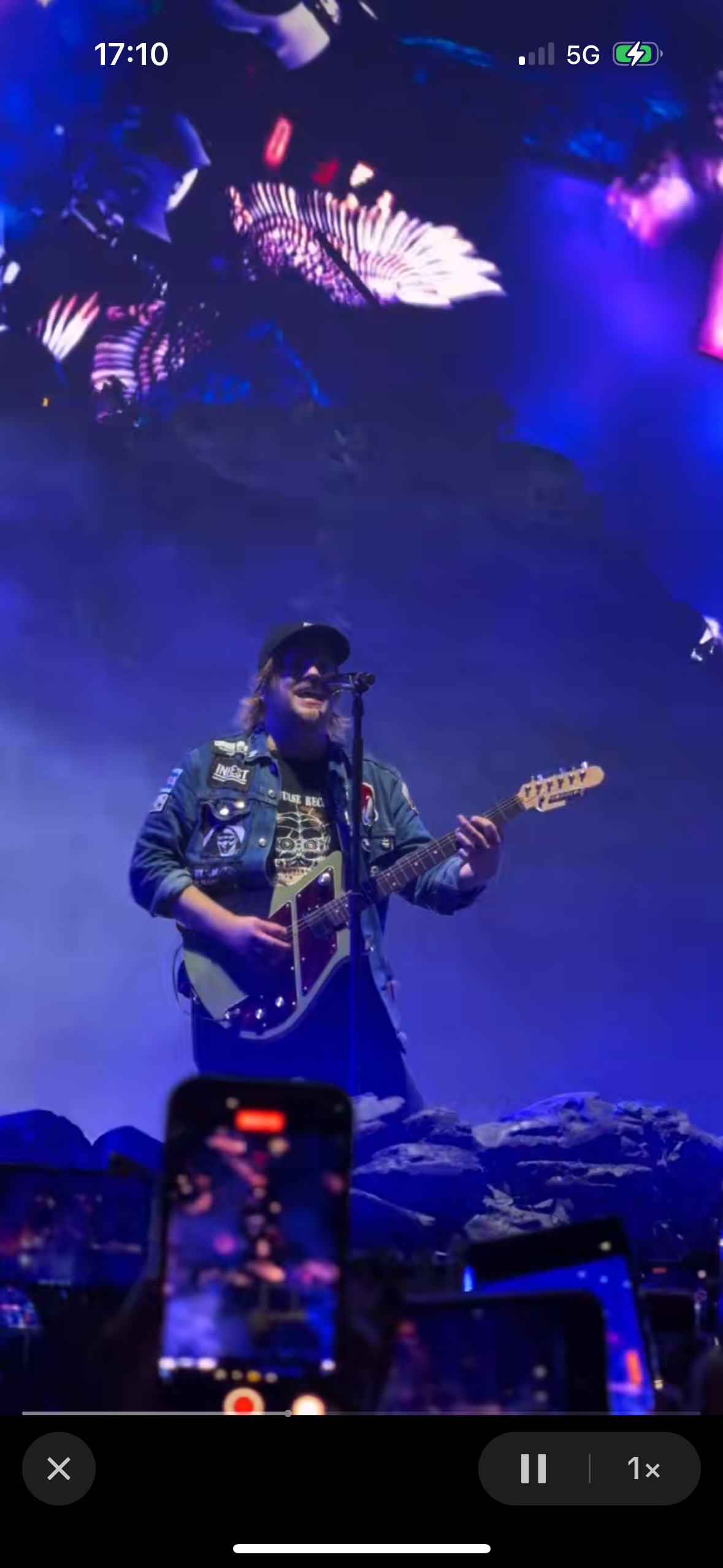 a musician on stage, holding a guitar and singing into a microphone. there are people in the background and the stage is dark with some lights on. the musician is wearing a jean jacket and is smiling.  the musician appears to be performing at a concert or festival.  the image is taken from a phone and the date and time is visible on the top of the image.  there is also a red dot visible on the phone's screen.