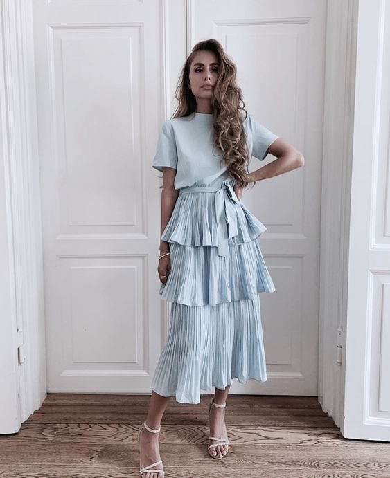 a fullbody shot of a woman with long blonde hair wearing a light blue tiered midi dress. the dress has a pleated skirt with three tiers and short sleeves. it is belted at the waist. she is wearing white heeled sandals and standing on a wooden floor in front of white doors. the overall style is elegant and summery.