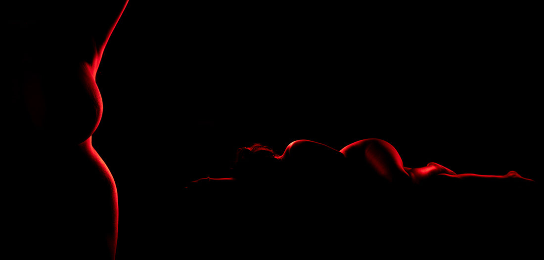 the image is a striking and artistic representation of two pregnant women's bodies. the left side shows a profile of a pregnant woman, primarily visible from the chest to hips. the right side depicts another pregnant woman lying down, her body curving into a graceful line across the frame.  both are rendered as silhouettes, illuminated by a vibrant red light that highlights the contours of their bodies. the dark background contrasts sharply with the red light, emphasizing the forms and creating a moody and sensual atmosphere.  the overall effect is abstract and evocative, focusing on the beauty of the female form during pregnancy.