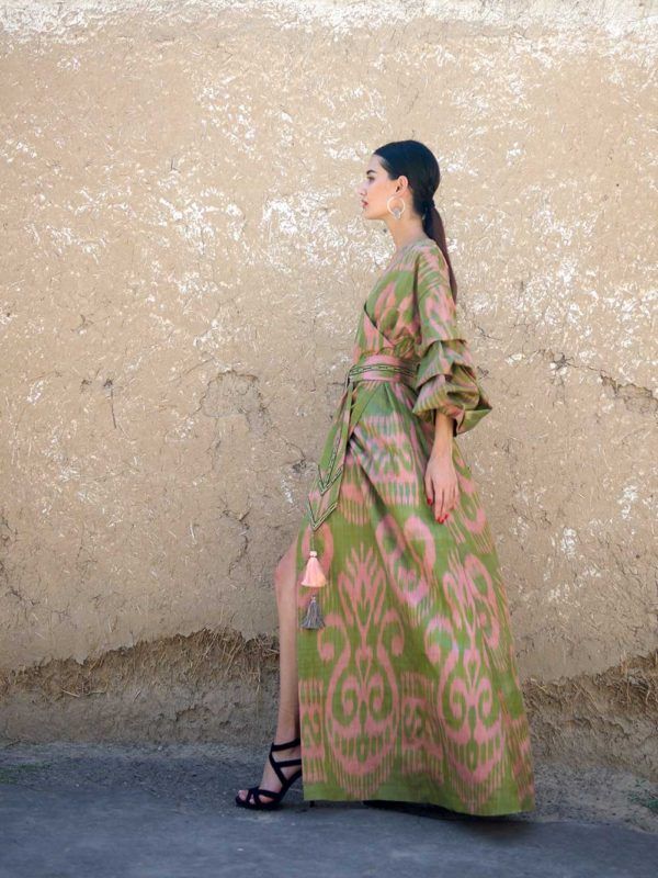 the image showcases a woman wearing a stunning maxi dress made from uzbek ikat fabric. the dress is predominantly green with a pink ikat pattern, featuring ruffled sleeves and a side slit. the model is posed against a textured, earthentoned wall, and her style is elegant and bohemian. she's accessorized with simple earrings and highheeled shoes. the overall setting and attire suggest a fashion shoot emphasizing the beauty and cultural significance of uzbek ikat textiles.