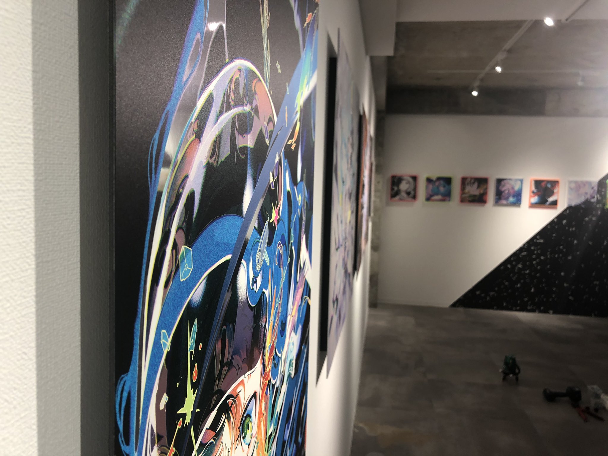 the image shows a series of colorful abstract paintings hanging on a white wall in an art gallery. the paintings are mounted on black canvases and feature a mix of bright colors and bold lines. the gallery is lit with artificial light, and the floor is a dark gray concrete.  the perspective is from eye level as if someone is walking through the space.