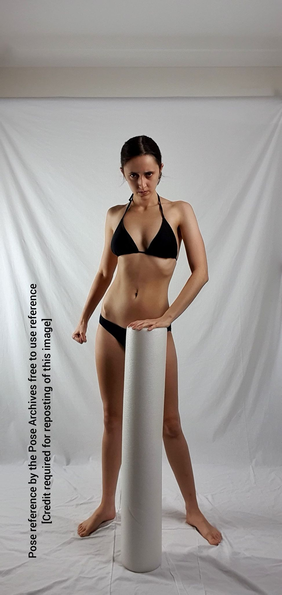 a young woman in a black bikini stands confidently, her left hand resting on a white pillar. the setting appears to be an indoor studio, with a white backdrop and soft, artificial lighting. the image is intended as a pose reference for artists.