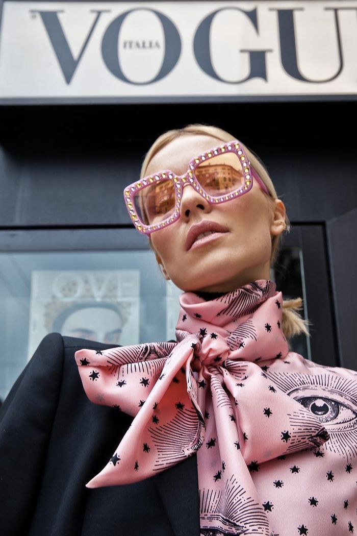 closeup portrait of a blonde woman wearing pink gucci sunglasses with studs, a pink silk scarf with an eye and star print, and a black blazer. the background features the vogue italia sign. the image is stylish and captures a highfashion street style aesthetic.