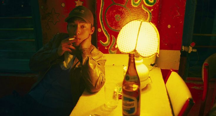 a man wearing a hat and jacket is sitting at a table with a beer bottle, glass, and lamp on it. he is smoking a cigarette. the background is a red wall with a design on it.