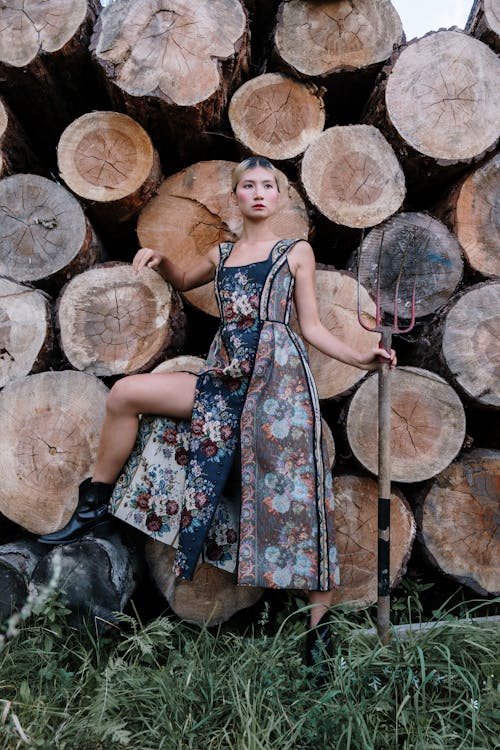 a full shot of a fashion model posing against a backdrop of stacked logs. she is wearing a long floral dress and black boots, and is holding a pitchfork. the setting is outdoors in a rural or countryside location. the image has a moody and mysterious atmosphere.
