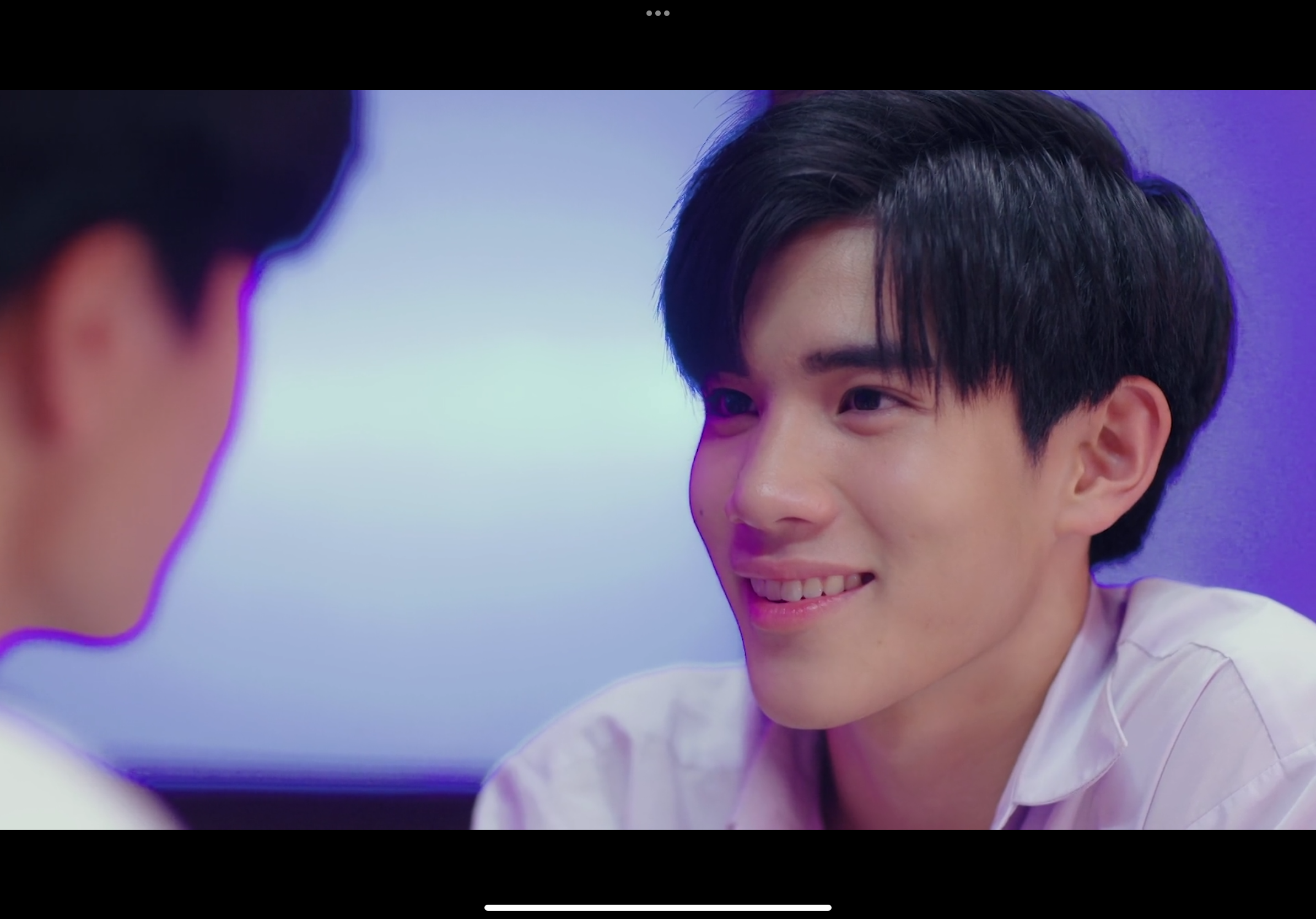 the image captures a closeup moment between two young men in a dimly lit, possibly school, setting. the focus is on the smiling face of the boy on the right, conveying a sense of warmth and happiness. the cooltoned lighting and blurred background emphasize the intimacy of the interaction. the image suggests a scene from a bl (boys' love) drama, hinting at a budding romantic connection.