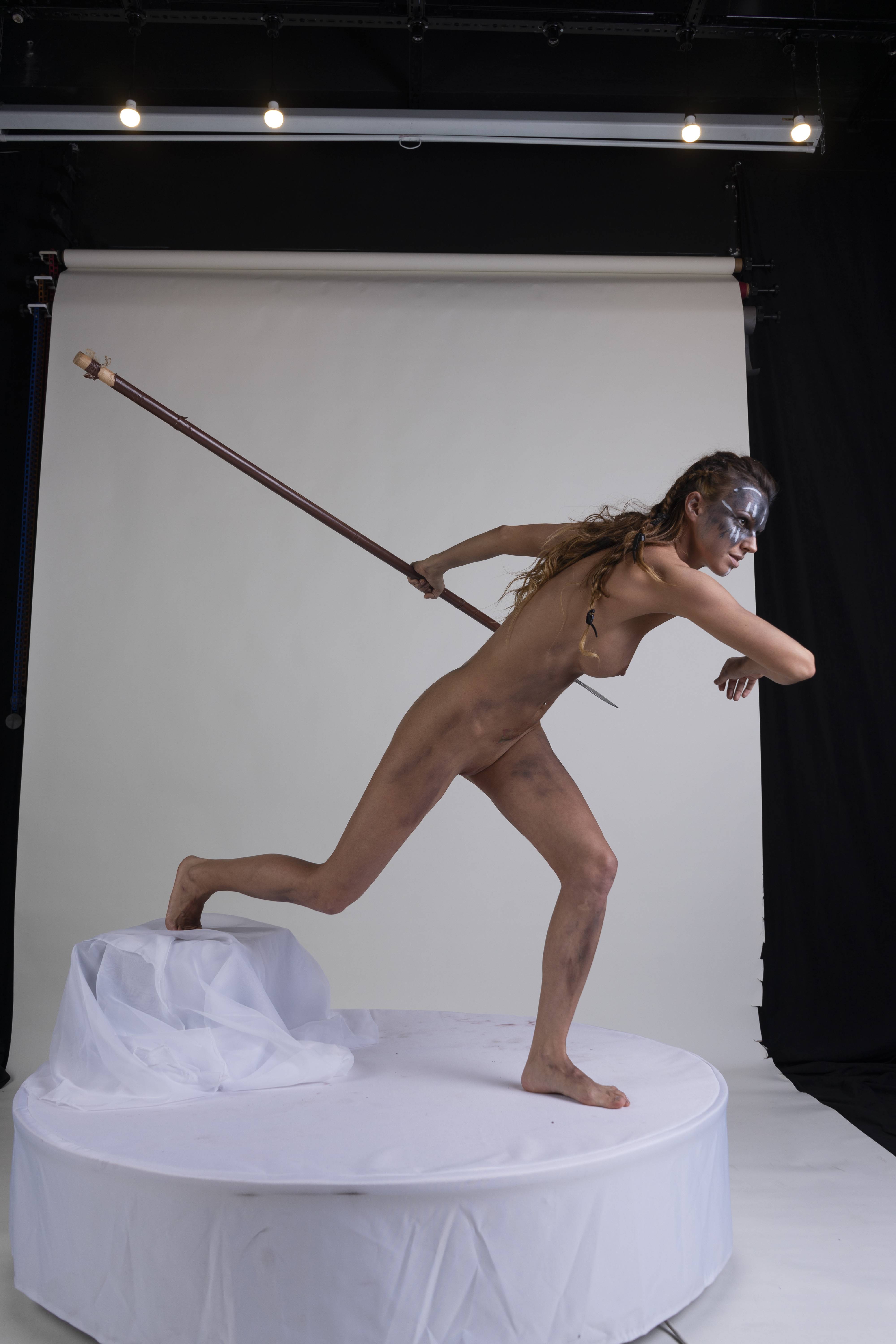 a nude woman with face paint is in an action pose with a spear in a studio setting. she has an athletic build and appears to be running on a white platform against a grey background. her pose suggests a warrior or mythical figure.