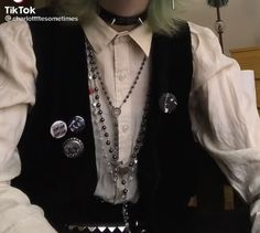 a young person wearing a white buttonup shirt with a black vest and chain necklaces. they are wearing a silver choker with spikes. the person is also wearing several pins on their vest, and is looking at the camera.