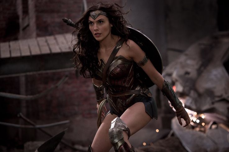 wonder woman, played by gal gadot, stands ready for battle, wielding her sword and shield. she's dressed in her iconic costume, with her hair flowing behind her. the scene is dark and gritty, suggesting an epic battle about to unfold. the image captures the power and intensity of wonder woman as a superhero and a warrior.