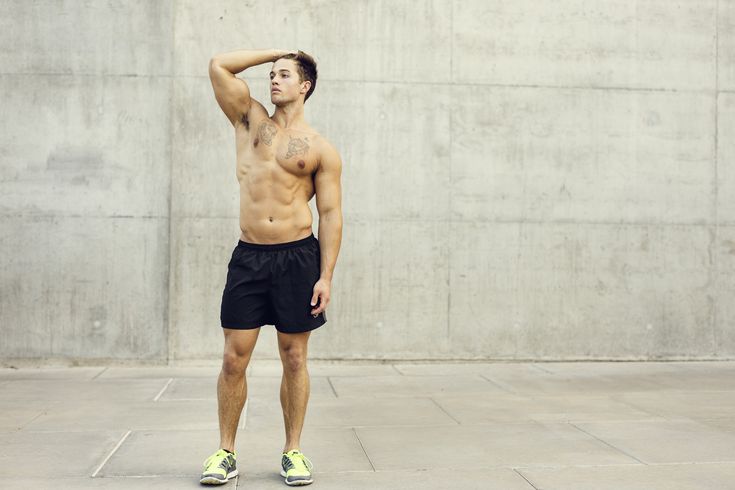 a muscular young man with tattoos on his chest is standing shirtless in black shorts and sneakers in front of a concrete wall. he is looking away from the camera and has his hand on his head.
