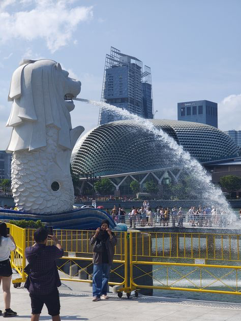 a wide shot of the merlion statue in singapore with a fountain spraying water into the air.  in the background are the esplanade  theatres on the bay and modern skyscrapers. tourists are visible along the waterfront, some taking photos. the scene is bright and sunny. a yellow temporary fence separates the pedestrian walkway from the water.