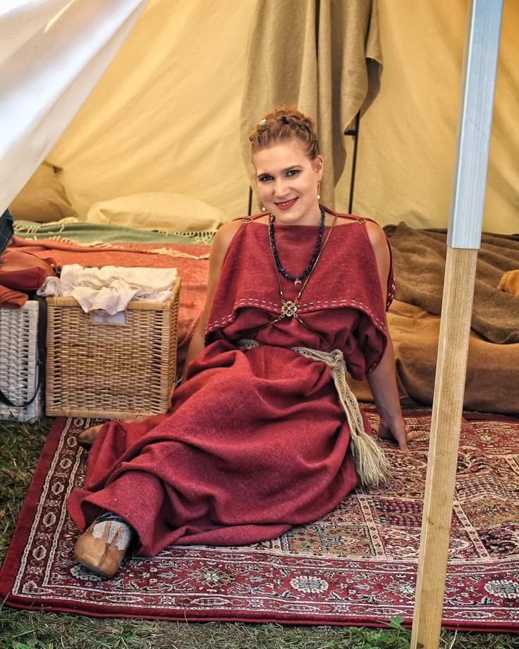 a woman wearing a red dress or tunic and jewelry is sitting inside a tent on a patterned rug. she is part of a historical reenactment. a basket is next to her, and pillows and blankets are visible inside the tent. the tent pole is visible on the right.