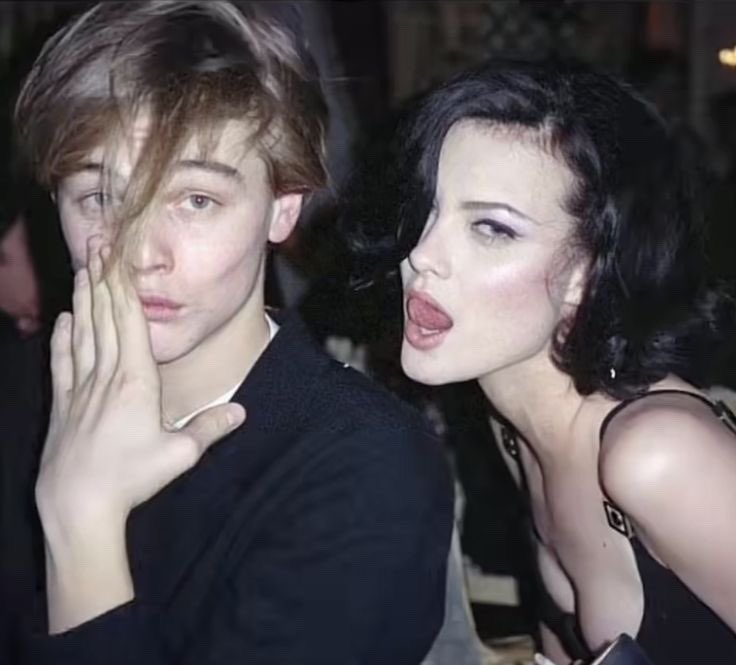 a vintage photograph shows leonardo dicaprio and liv tyler at a public event. dicaprio, with brown hair partially covering his face, is holding his hand up near his face. tyler, with dark hair and a black dress, is standing beside him with her tongue slightly out.