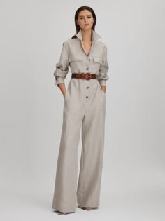a woman is wearing a beige linen jumpsuit with a wide leg and long sleeves. she is standing in a studio, with a white background. the jumpsuit has a belt around the waist and is buttoned up the front. the woman is wearing a casual and elegant look.