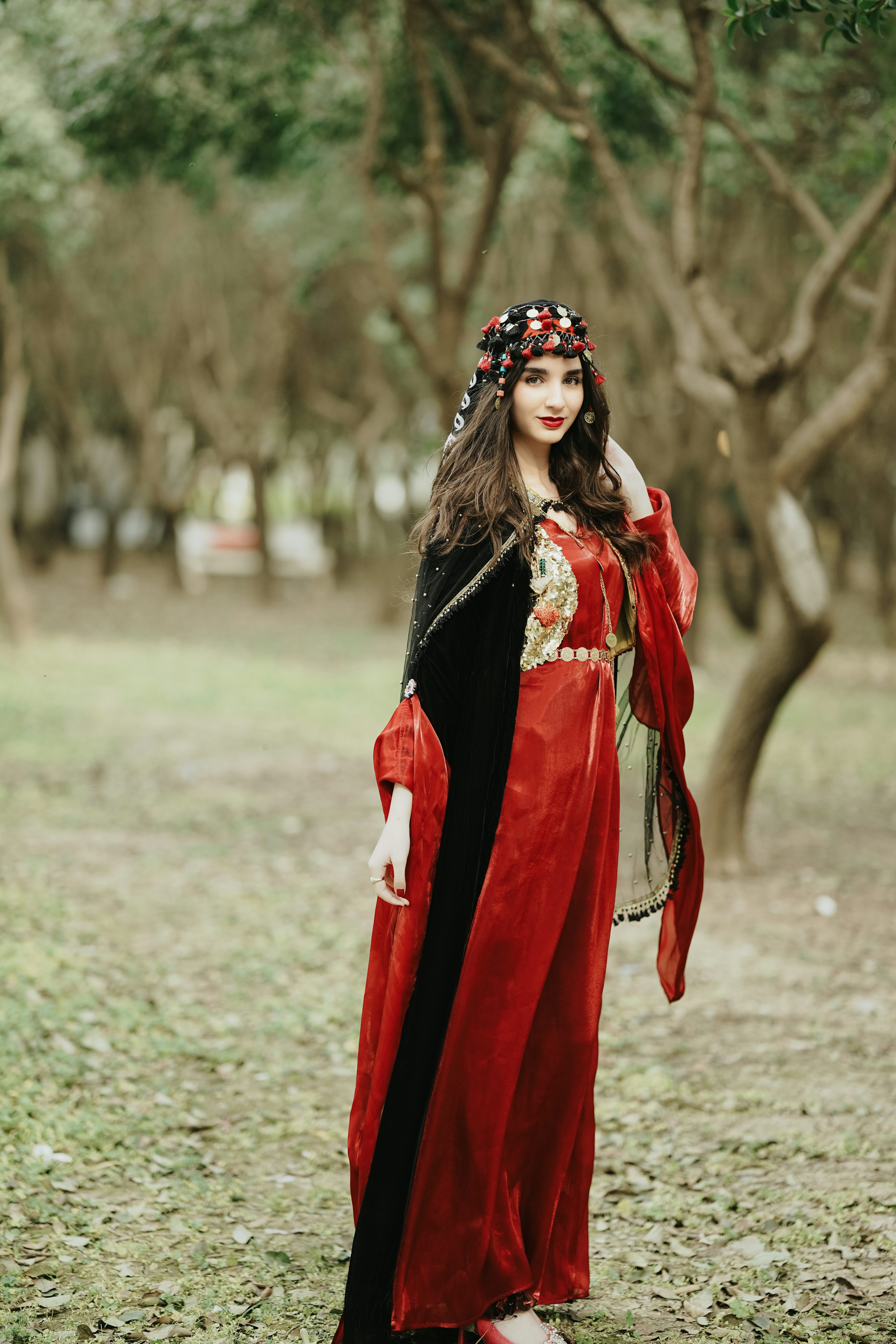 a woman stands in a forest wearing traditional red and black clothing with a headdress. she is positioned in the center of the frame, looking towards the camera. the background consists of trees and foliage.