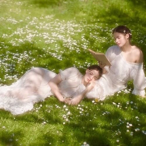 two women in white dresses lie on a grassy field dotted with daisies. one woman is asleep, resting her head on the lap of the other, who is sitting up and reading a book. the scene is bathed in soft, ethereal light, creating a peaceful and idyllic atmosphere.