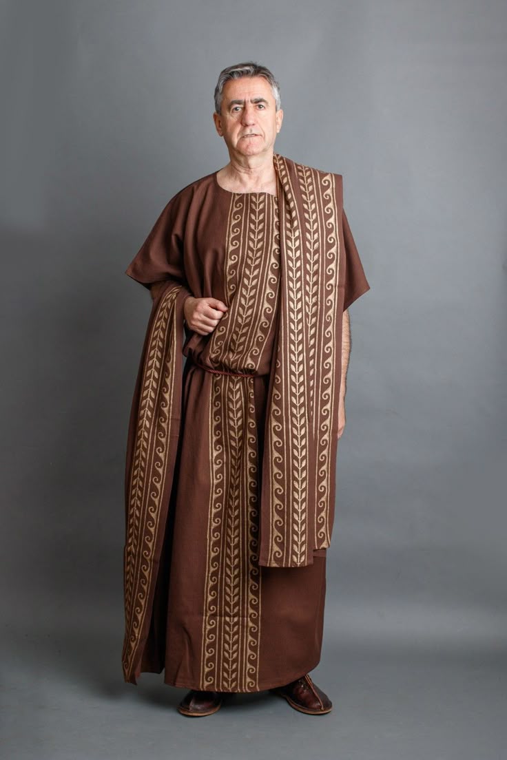 a full shot of a middleaged man standing against a gray background. he is dressed in a brown toga, adorned with golden wave and leaf patterns on the vertical edges and across the shoulder piece. the toga is secured at the waist with a cord. he is wearing brown shoes. the man has short gray hair, and a serious expression.