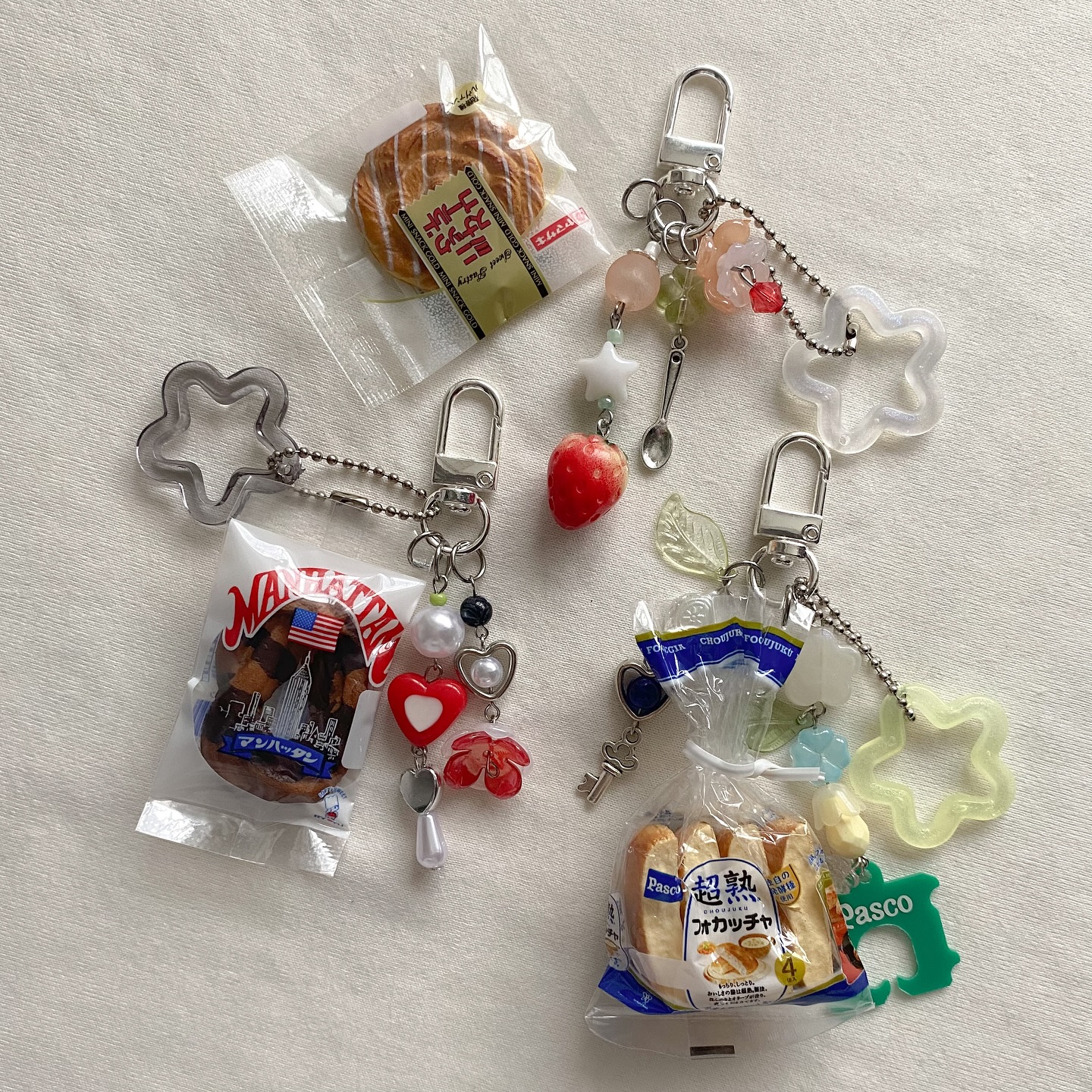 the image shows a collection of four unique foodthemed keychains. each keychain features a realistic miniature of different types of bread and pastries, sealed in plastic bags and enhanced with various charms. the top left keychain shows a wrapped pastry, the bottom left is a manhattan bakery item with an american flag, the top right has an apple charm, and the bottom right has a pasco focaccia with a green dog charm. the keychains are attached to silver clasps and chains, adorned with beads, hearts, spoons, stars, and other decorative elements, creating a visually appealing and detailed collection of novelty accessories.
