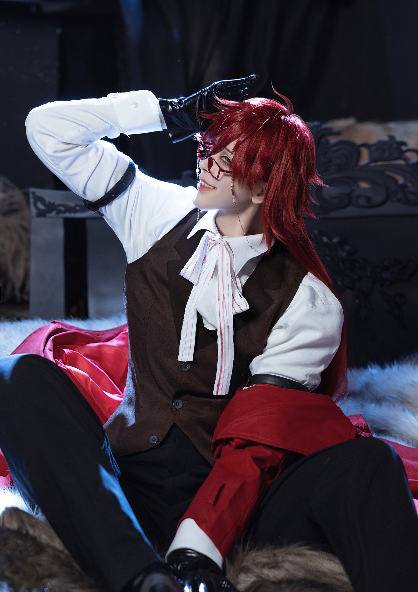 a cosplayer, dressed as an anime character, sits on a fluffy white rug. the cosplayer is male with long red hair, red and black clothing, black gloves, and glasses. he is smiling and looking toward the right of the frame, as he holds his hand to his head.