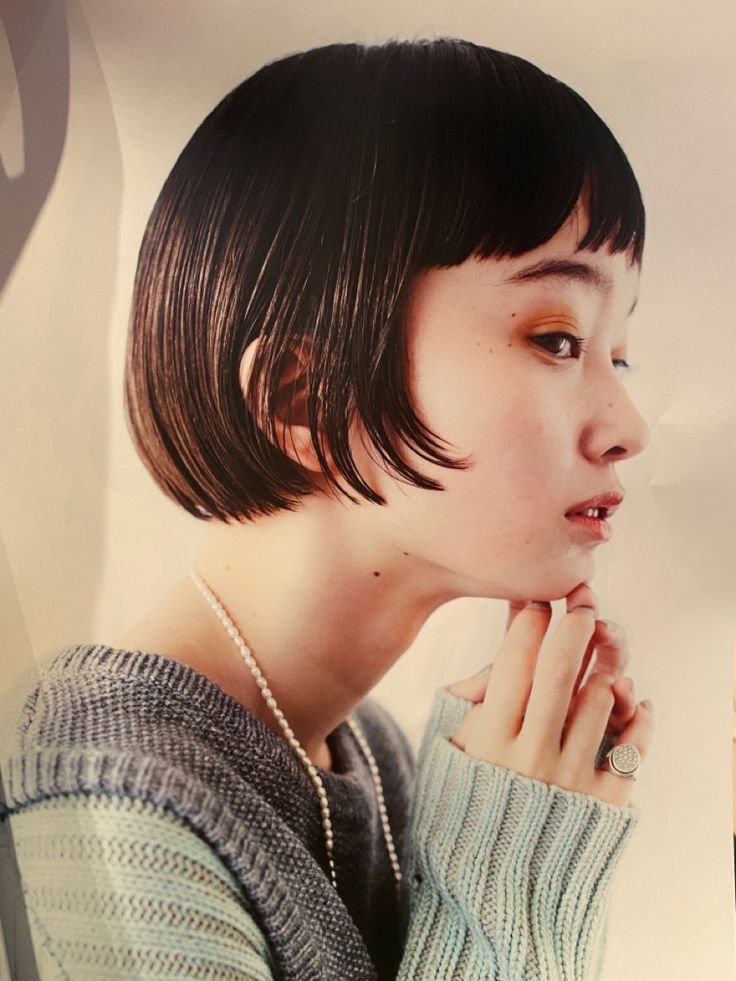 a woman with short black bob hair, wearing a grey and blue sweater, is looking away from the camera, and her chin is resting on her hand. she is wearing a pearl necklace and a ring on her finger. the background is white.