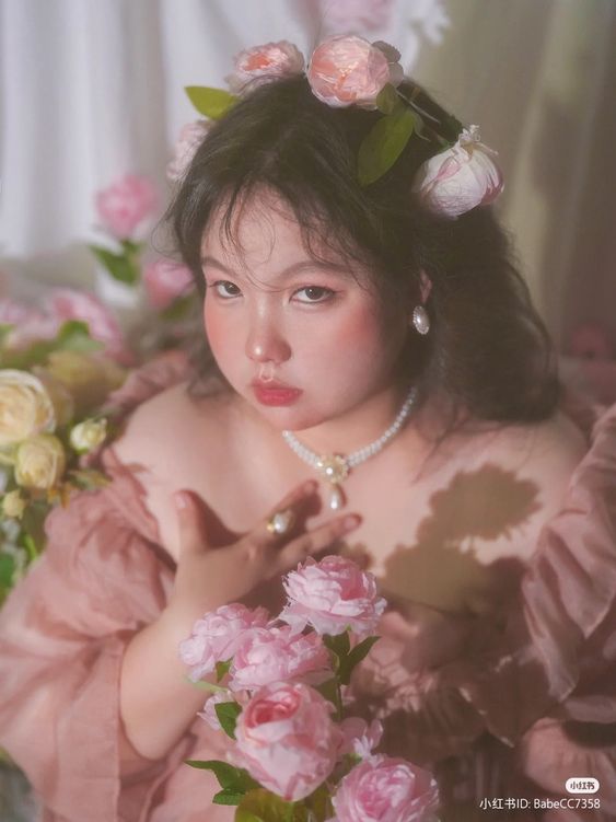 a young asian girl with a cherubic face and rosy cheeks is the focal point of this portrait. she is dressed in a soft, peachcolored offtheshoulder dress with a ruffled neckline. delicate pink roses adorn her hair in a crownlike arrangement, and she wears a pearl necklace and ring, adding to the overall sense of sweetness and innocence. her expression is serious yet captivating, her dark eyes looking directly at the viewer. the background is a blur of light colors, further emphasizing the girl as the central element. the lighting is soft and diffused, casting a gentle glow on the subject and creating a dreamy atmosphere. the overall composition and color palette contribute to a sense of tenderness and childlike beauty.