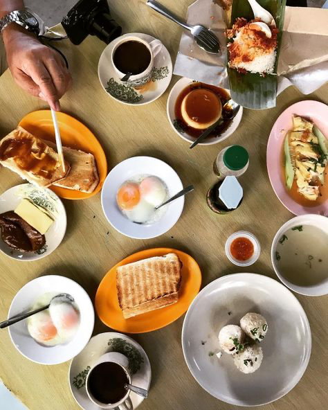 a table full of food, including toast, eggs, coffee, tea, rice, chicken, and dessert. the food appears to be asian in origin, possibly singaporean or malaysian. the table is set with a variety of plates and bowls, and there are napkins and utensils available. the food looks delicious and inviting.