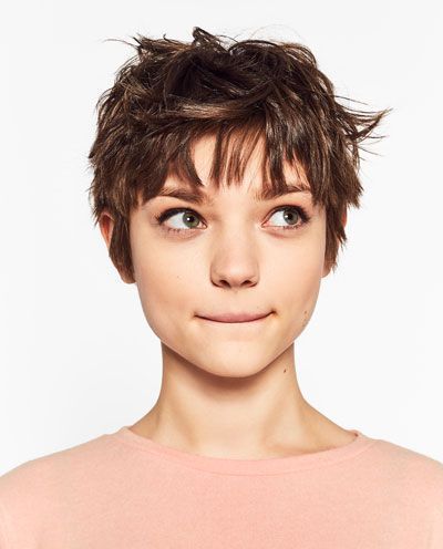 a young woman with short brown hair in a pixie cut style is shown in a portrait against a white background. she is wearing a pale pink tshirt and is looking slightly up and to the left with a curious expression. the image is welllit with studio lighting, creating a clean and bright aesthetic.