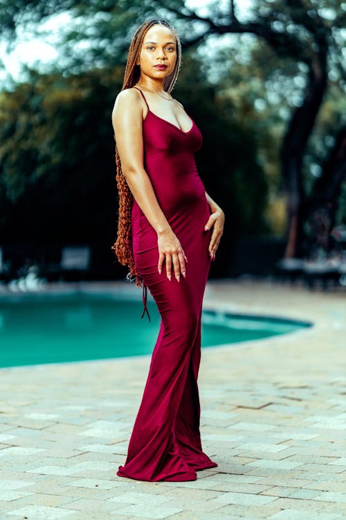 a fulllength photograph showcases a woman of color, confidently posing poolside in a formfitting, floorlength maroon dress. her dark brown hair is styled in long braids which fall down past her shoulders. the setting is outdoors, with lush green foliage slightly blurred in the background, implying a garden or resort environment. the overall ambiance evokes a summery, elegant mood. the woman's pose is natural, and her expression is both composed and alluring. the dress is a key element, and its rich color contrasts nicely against her skin tone.  the pool in the background is a teal color. the paving stones where she stands are a lightcolored, neutral stone.