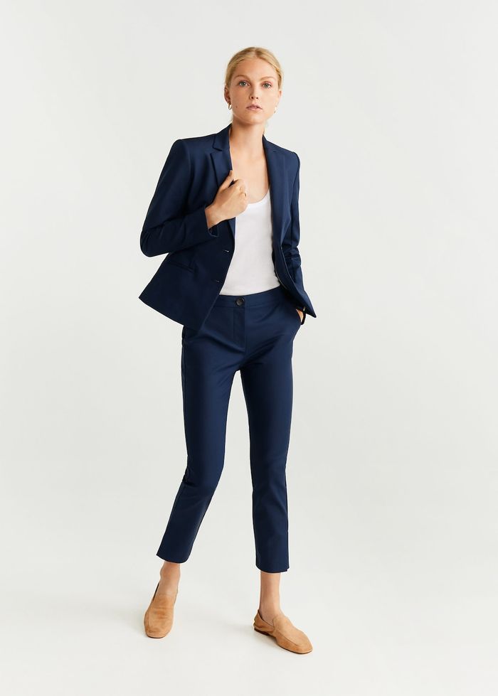 a fullbody studio shot of a woman model wearing a navy blue suit. the suit consists of a blazer and cropped trousers. she is also wearing tan loafers. the background is plain white. the overall style is business casual and elegant.