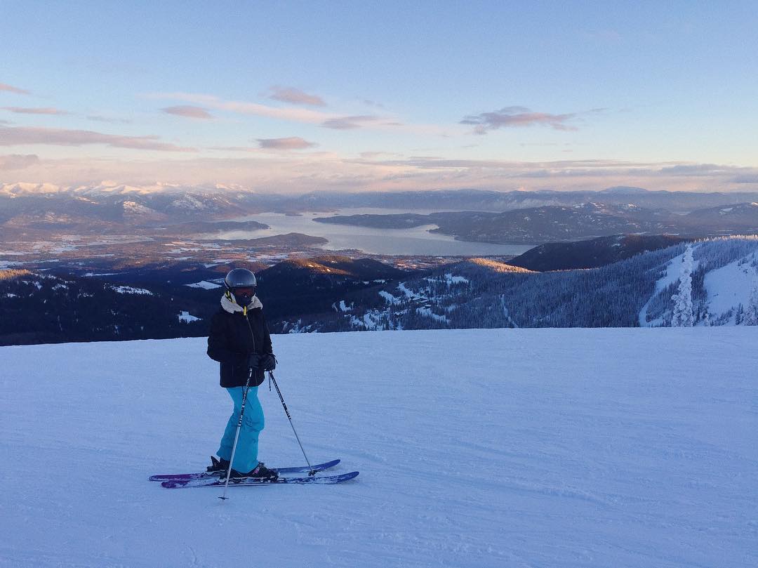 a lone skier stands on a snowy mountain peak, gazing out at a breathtaking panoramic view of a valley, lake, and distant mountain ranges. the sun is setting, casting a warm glow over the landscape. the skier is dressed in warm winter gear, and their skis are planted firmly in the snow. the scene is peaceful, serene, and evokes a sense of adventure and freedom.